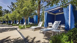 A row of outdoor lounge chairs under blue canopies, with palm trees and greenery around.