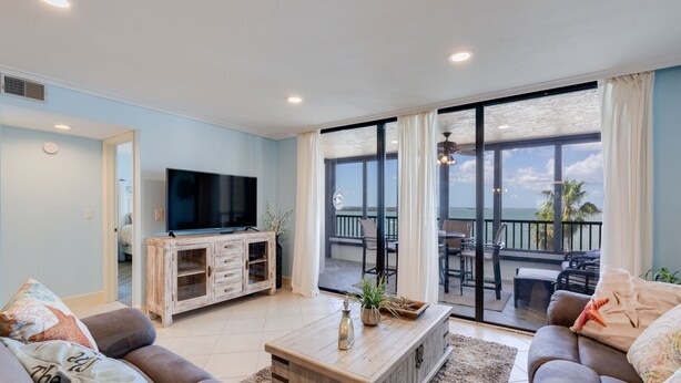 A living room with a TV, wooden cabinet, and a large sliding glass door leading to a balcony with outdoor furniture. - Gorgeous Punta Rassa Condo 504 - River and Sunset views! (Fort Myers)