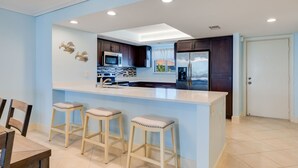 A modern kitchen with a central island, bar stools, and a dining area with chairs. - Gorgeous Punta Rassa Condo 504 - River and Sunset views! (Fort Myers)