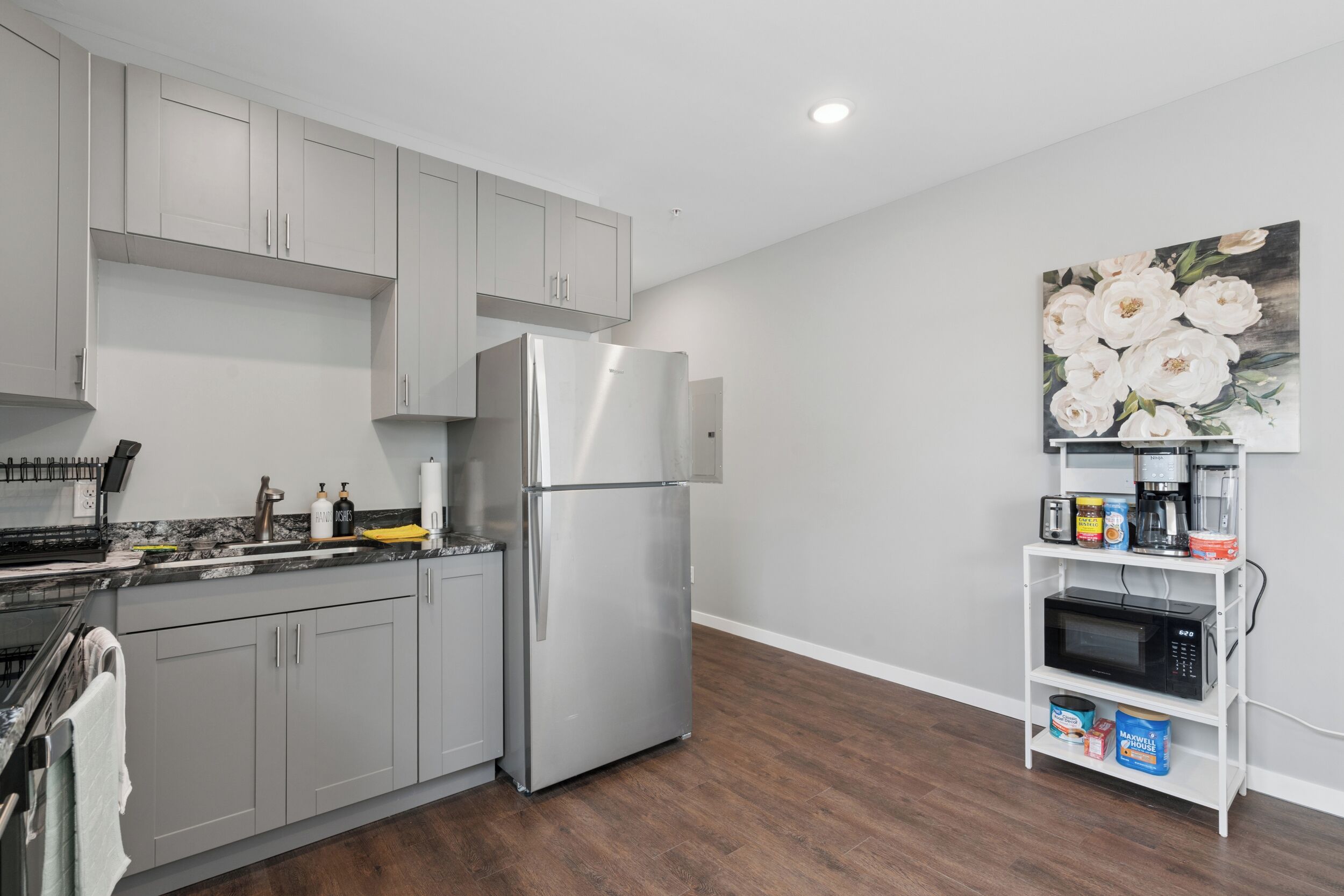 A modern kitchen with grey cabinets, a stainless steel refrigerator, a microwave, and a shelf with various items.