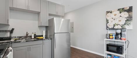A modern kitchen with grey cabinets, a stainless steel refrigerator, a microwave, and a shelf with various items.