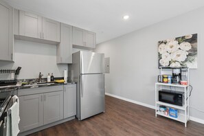 A modern kitchen with grey cabinets, a stainless steel refrigerator, a microwave, and a shelf with various items. - Valclyde Cozy Suite-Sleeps 4-Downtown-MPLs (Minneapolis)