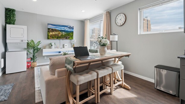 A modern kitchen with a refrigerator, a microwave, and a sink. There is a dining area with a table, chairs, and a laptop. A large window provides a view of the city. - Valclyde Cozy Suite-Sleeps 4-Downtown-MPLs (Minneapolis)