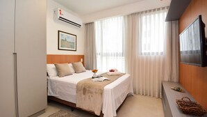 A hotel room with a bed, a TV, a window with curtains, an air conditioner, and a framed picture on the wall. - Studio em frente a UFSC e HU - 106A VIVA (Florianópolis)