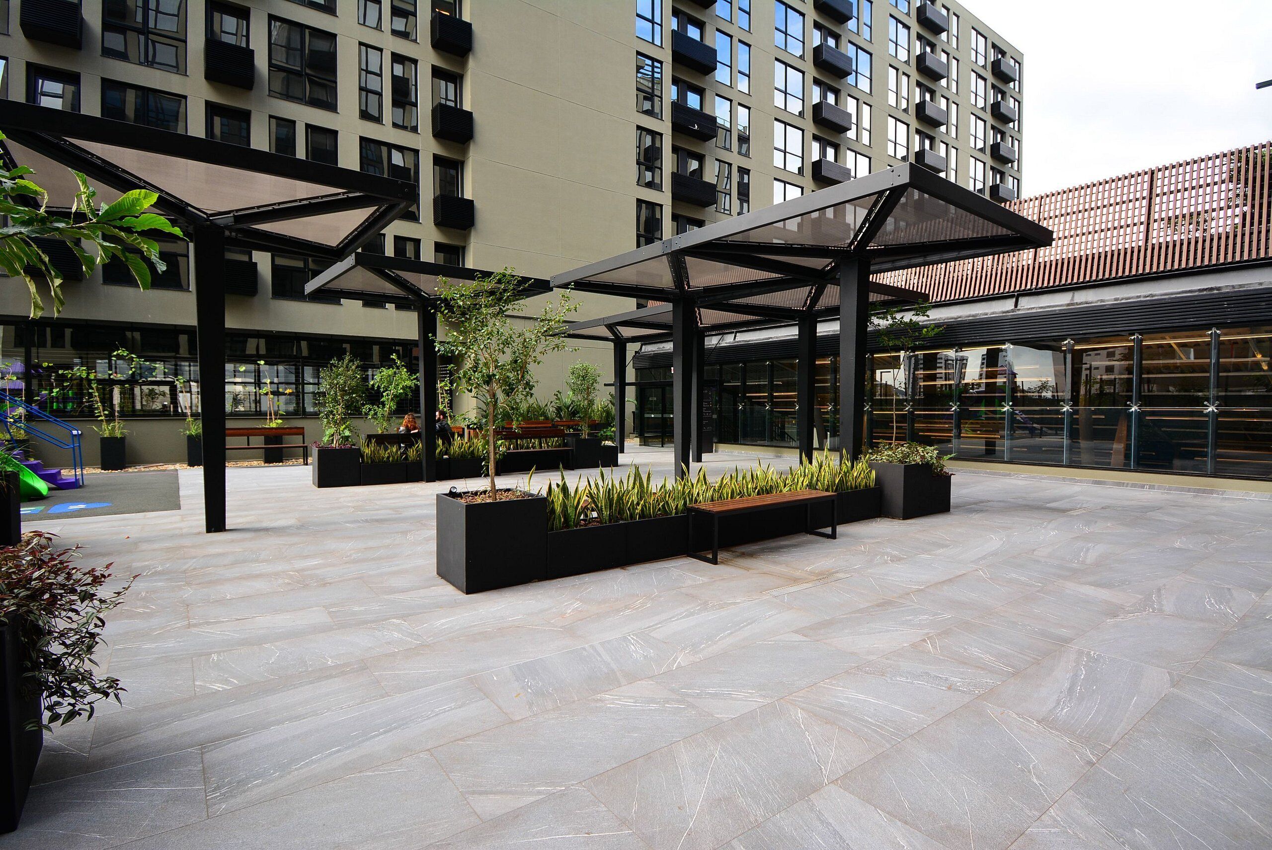 An outdoor courtyard with modern architectural design, featuring geometric patterns on the floor, black planters with greenery, and a covered seating area.