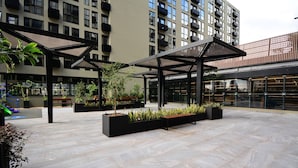 An outdoor courtyard with modern architectural design, featuring geometric patterns on the floor, black planters with greenery, and a covered seating area. - Studio em frente a UFSC e HU - 106A VIVA (Florianópolis)