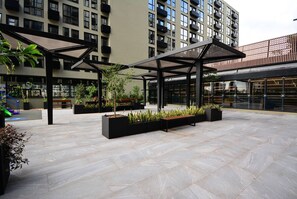 An outdoor courtyard with modern architectural design, featuring geometric patterns on the floor, black planters with greenery, and a covered seating area. - Studio em frente a UFSC e HU - 106A VIVA (Florianópolis)