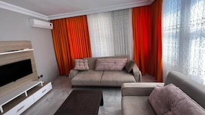 A modern living room with a sofa, a TV, and a large window with orange curtains.