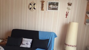 Un letto con una testiera nera, una coperta blu e un cuscino con un motivo a uccello. C'è una sveglia da parete con la scritta 'BIENVENUE A BORD', un quadro incorniciato raffigurante una barca e una lampada rossa.