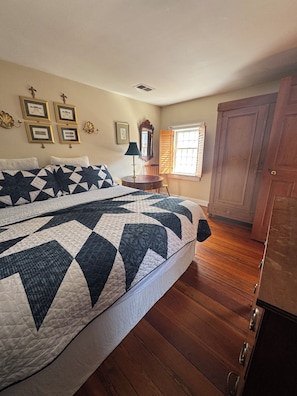A bedroom with a large bed, wooden flooring, a dresser, a window with blinds, and framed pictures on the wall.