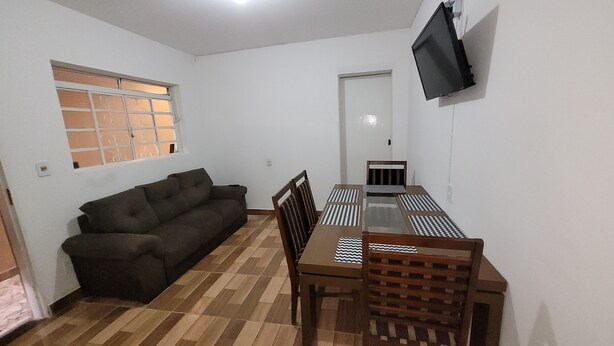 Una habitación con un sofá, una mesa de comedor con sillas, un televisor montado en la pared y una ventana.