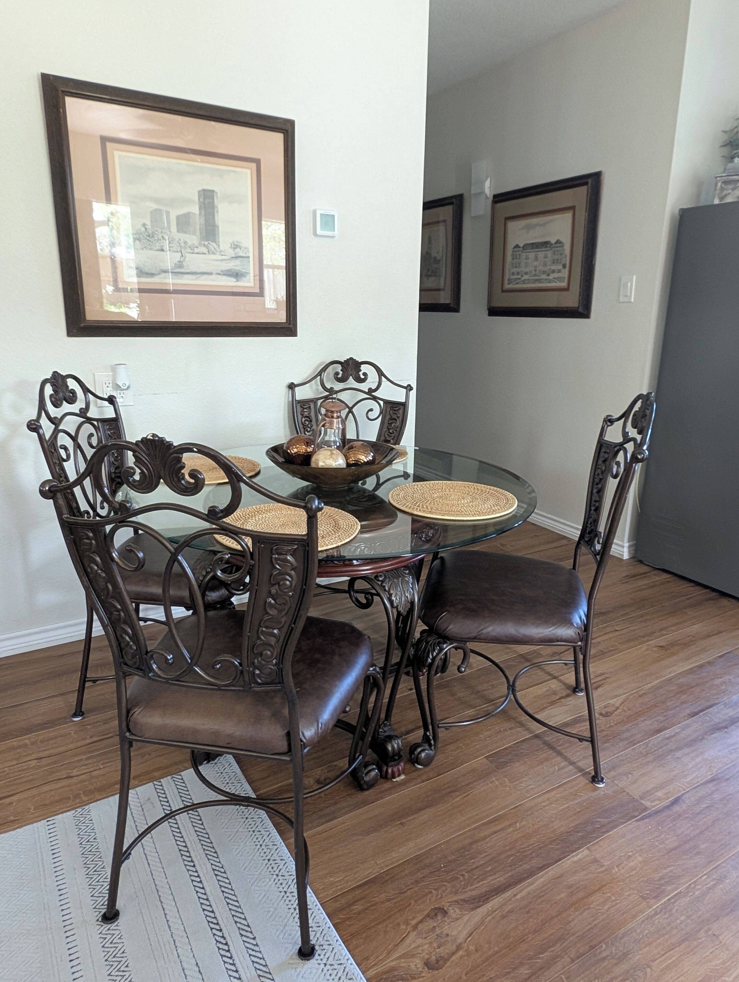 A dining area with a round glass table, four dark brown chairs with intricate designs, a woven placemat, and a framed picture on the wall.