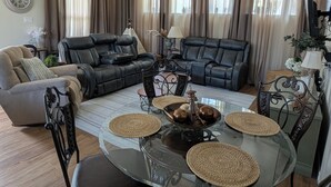A living room with a glass coffee table, two black leather sofas, a beige armchair, and a dining table with woven placemats.