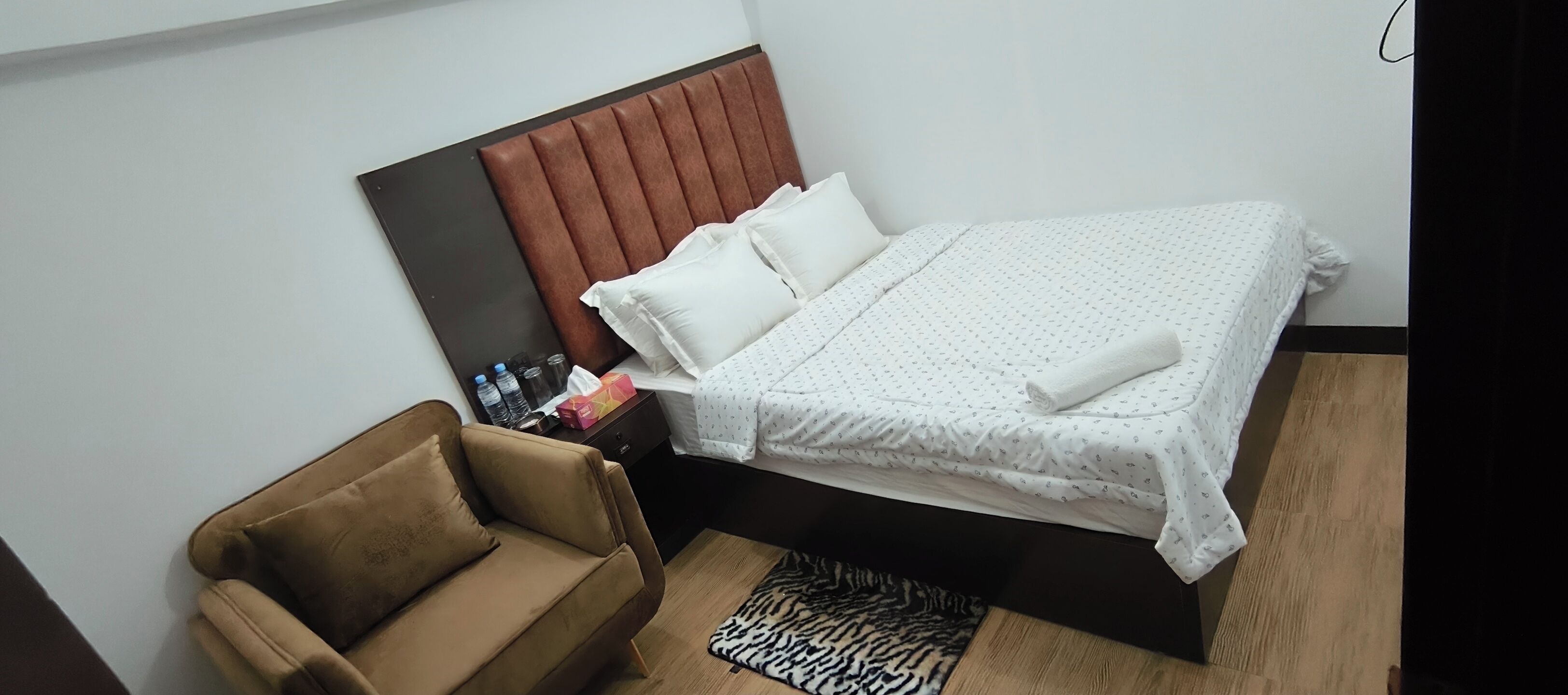 A hotel room with a bed, a brown sofa, a nightstand, and a zebra-patterned rug.