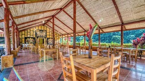 An open-air dining area with wooden tables and chairs, a tiled floor, and a thatched roof. - Villa 9B Toucan Valley Jungle Village Resort (Ciudad Cortés)