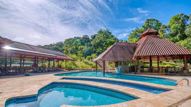 A swimming pool with a curved design, surrounded by stone tiles. There are covered seating areas with tables and chairs. - Villa 9B Toucan Valley Jungle Village Resort (Ciudad Cortés)