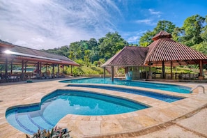 A swimming pool with a curved design, surrounded by stone tiles. There are covered seating areas with tables and chairs. - Villa 9B Toucan Valley Jungle Village Resort (Ciudad Cortés)