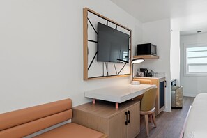 Ein Hotelzimmer mit einem Bett, einem Schreibtisch mit Stuhl, einem Fernseher, einer Lampe und einer kleinen Küchenzeile.
