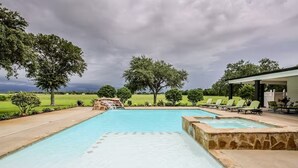 A rectangular swimming pool with a stone border, surrounded by a patio area with lounge chairs and a covered seating area.