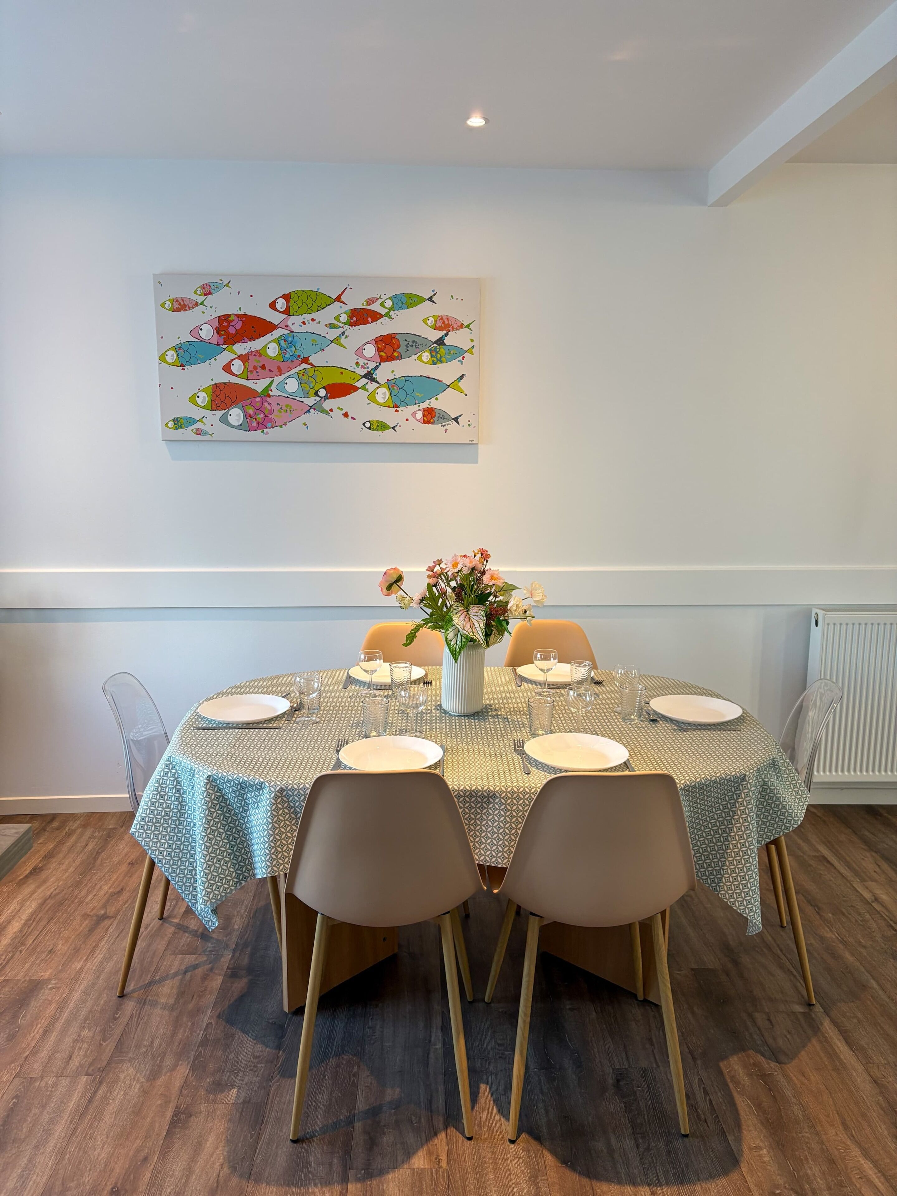 A dining area with a table set for four, clear chairs, a vase with flowers, and a colorful fish painting on the wall.