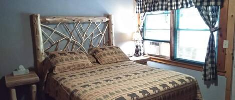 Una cama con colcha estampada, cabecera de madera, mesita de noche con lámpara y ventana con cortinas.