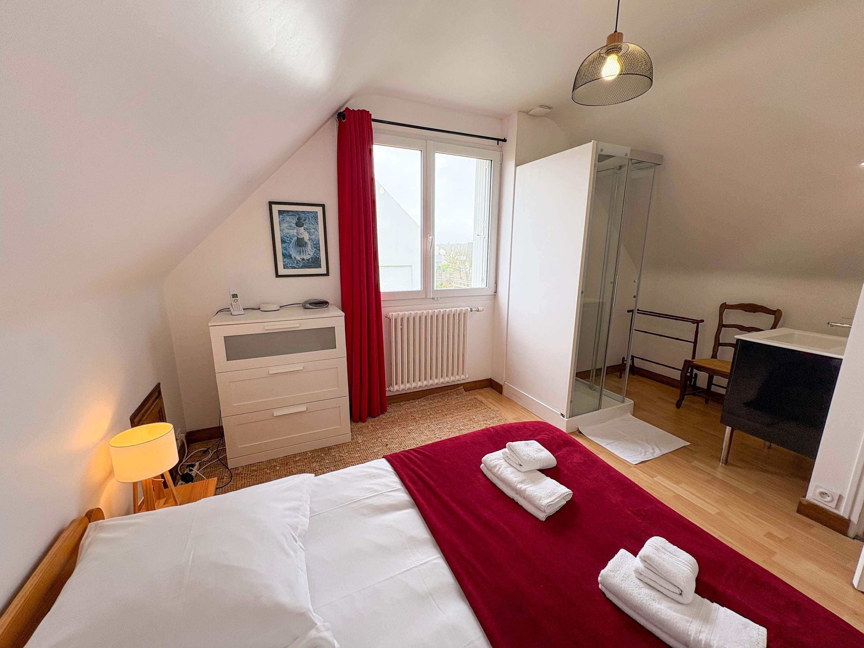A bedroom with a bed, a desk, a chair, a radiator, a shower, and a window with curtains.