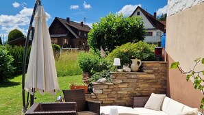 Een terras buiten met rieten meubels, een stenen muur en een witte parasol.