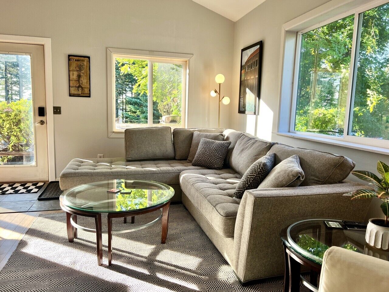 A modern living room with a sectional sofa, a glass coffee table, and a view of greenery outside.