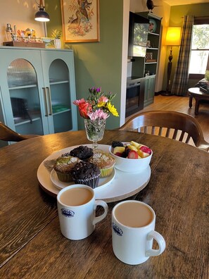 Una mesa con una bandeja de muffins, un bol de fruta y dos tazas de café.