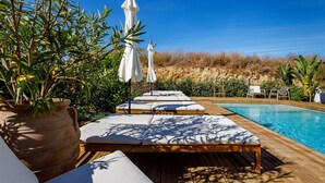 Zona junto a la piscina con sillas de descanso y sombrillas, una terraza de madera y un cielo azul despejado.