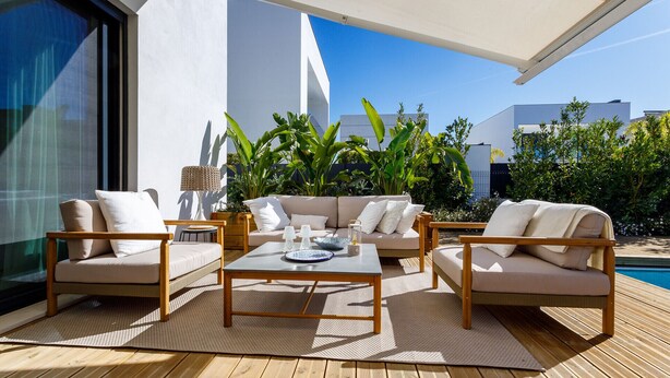 Un área de estar al aire libre con sofá, sillas y una mesa de centro, rodeada de plantas y una piscina.