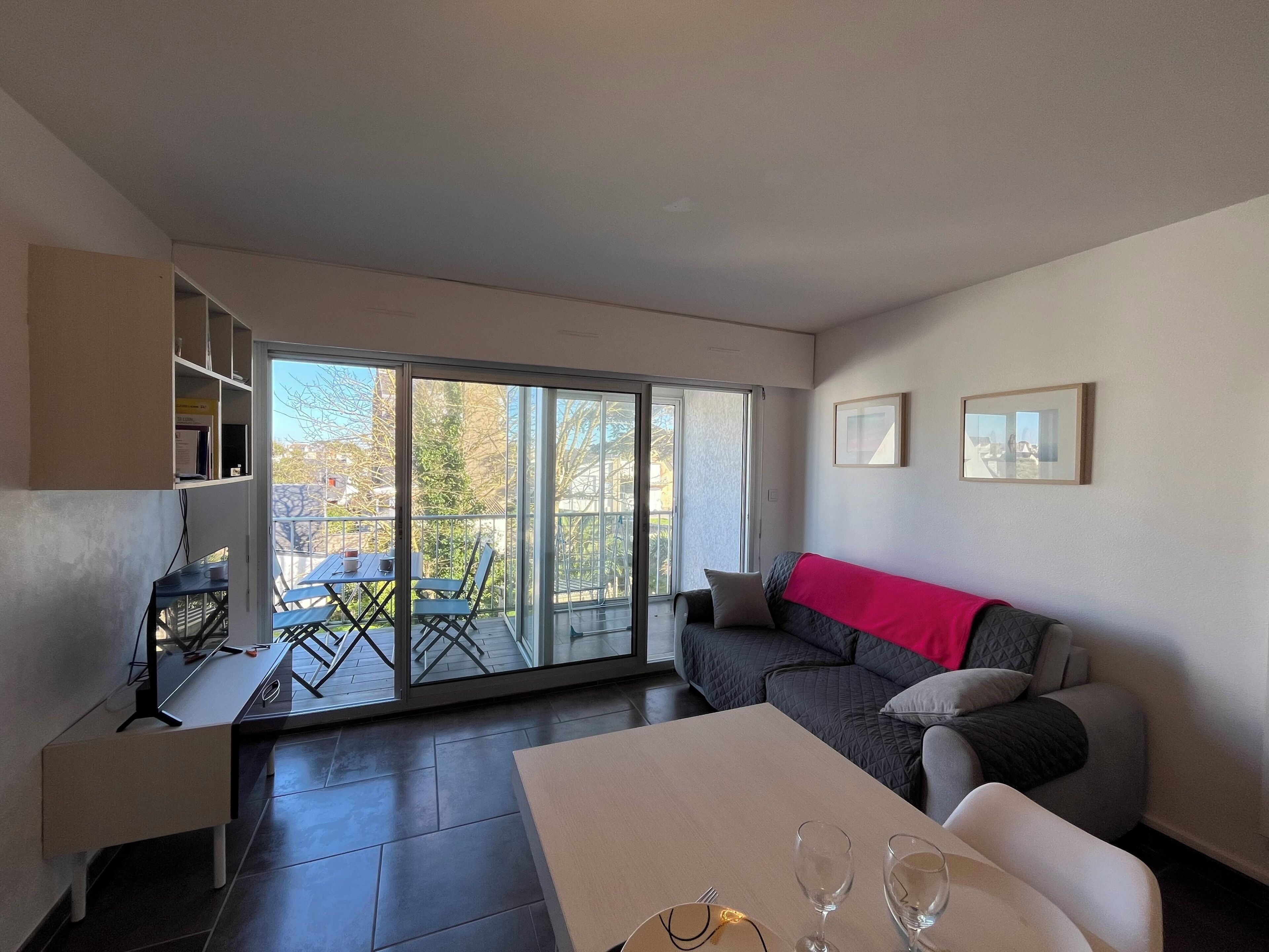 A modern living room with a sofa, a dining table with wine glasses, a balcony with a table and chairs, and a sliding glass door leading outside.