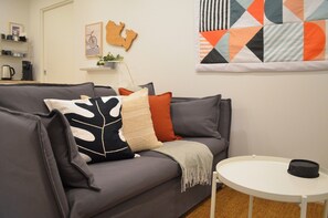 A modern living room with a grey sofa, a small white table, and a colorful quilt on the wall. - Skovhaven West Coast Suite in Upper Gibsons, minutes away from the beach & more! (Gibsons)