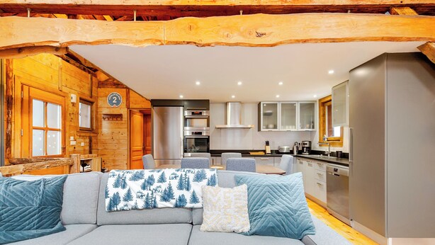 Un interior de cabaña rústica moderna con un sofá gris, paredes de madera, una cocina con electrodomésticos de acero inoxidable y un comedor.