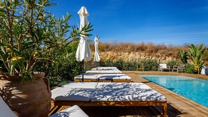 Zona junto a la piscina con sillas de descanso y sombrillas, una terraza de madera y un cielo azul despejado.