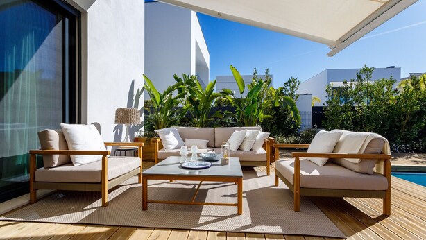 Un área de estar al aire libre con sofá, sillas y una mesa de centro, rodeada de plantas y una piscina.