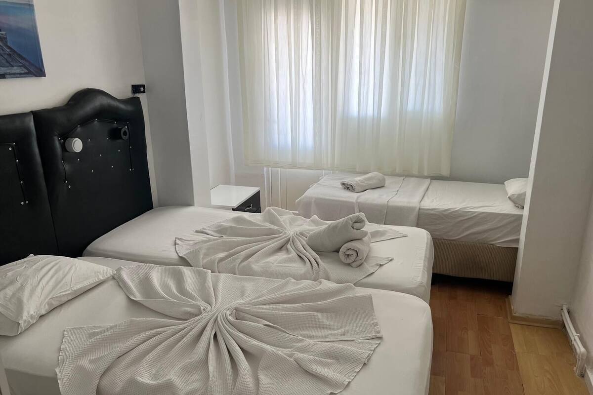 Quarto de hotel com duas camas, cabeceira preta, mesa de cabeceira e janela com cortinas leves.