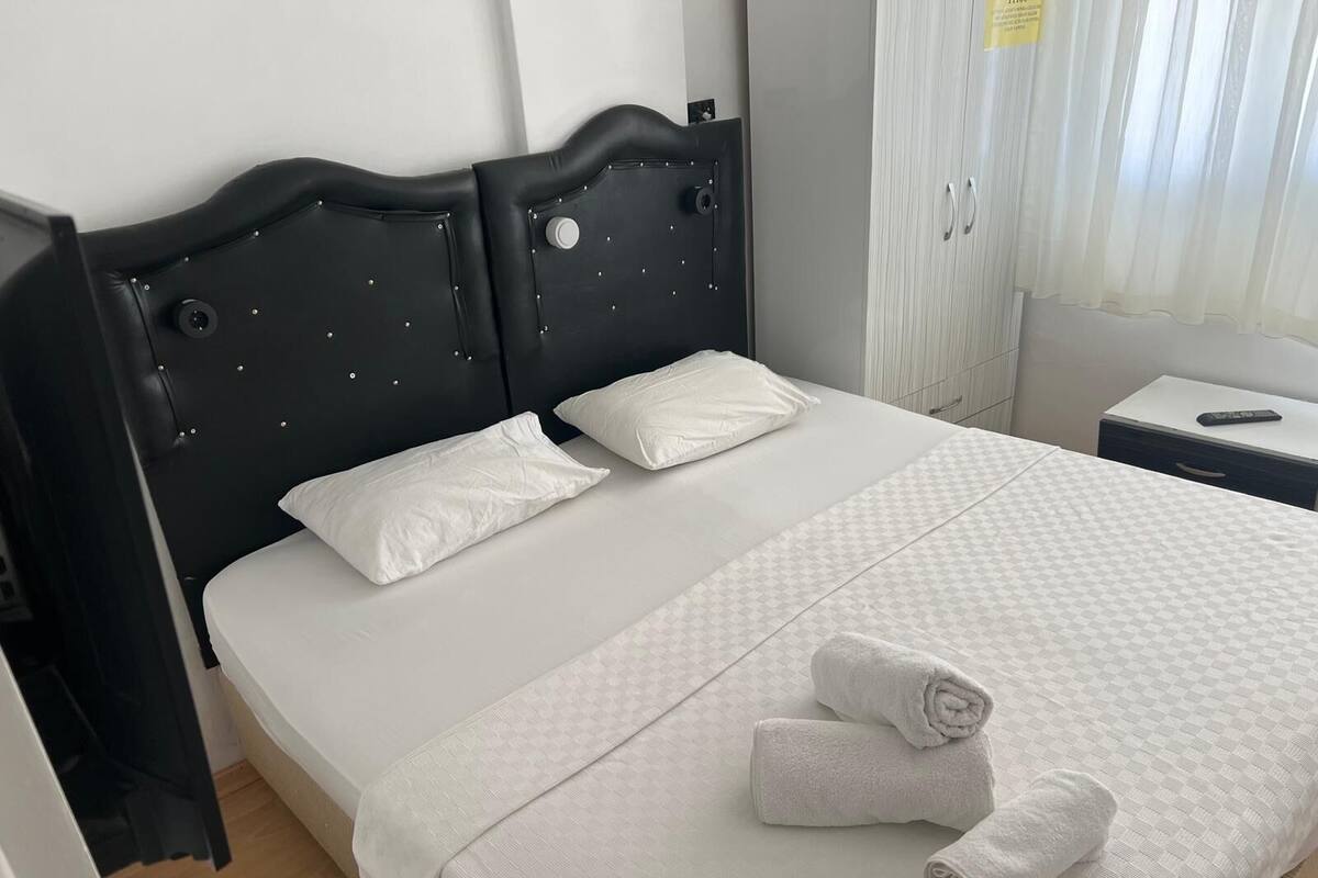 Cama bem arrumada com lençóis brancos e duas almofadas, cabeceira preta, mesa de cabeceira com controle remoto e janela com cortinas finas.