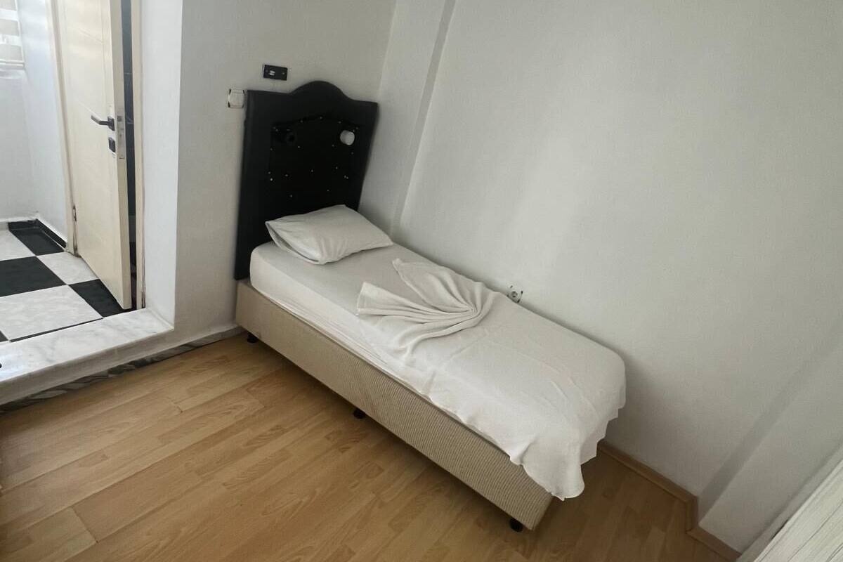 Quarto pequeno com uma cama de solteiro, piso de madeira, porta branca e espelho na parede.