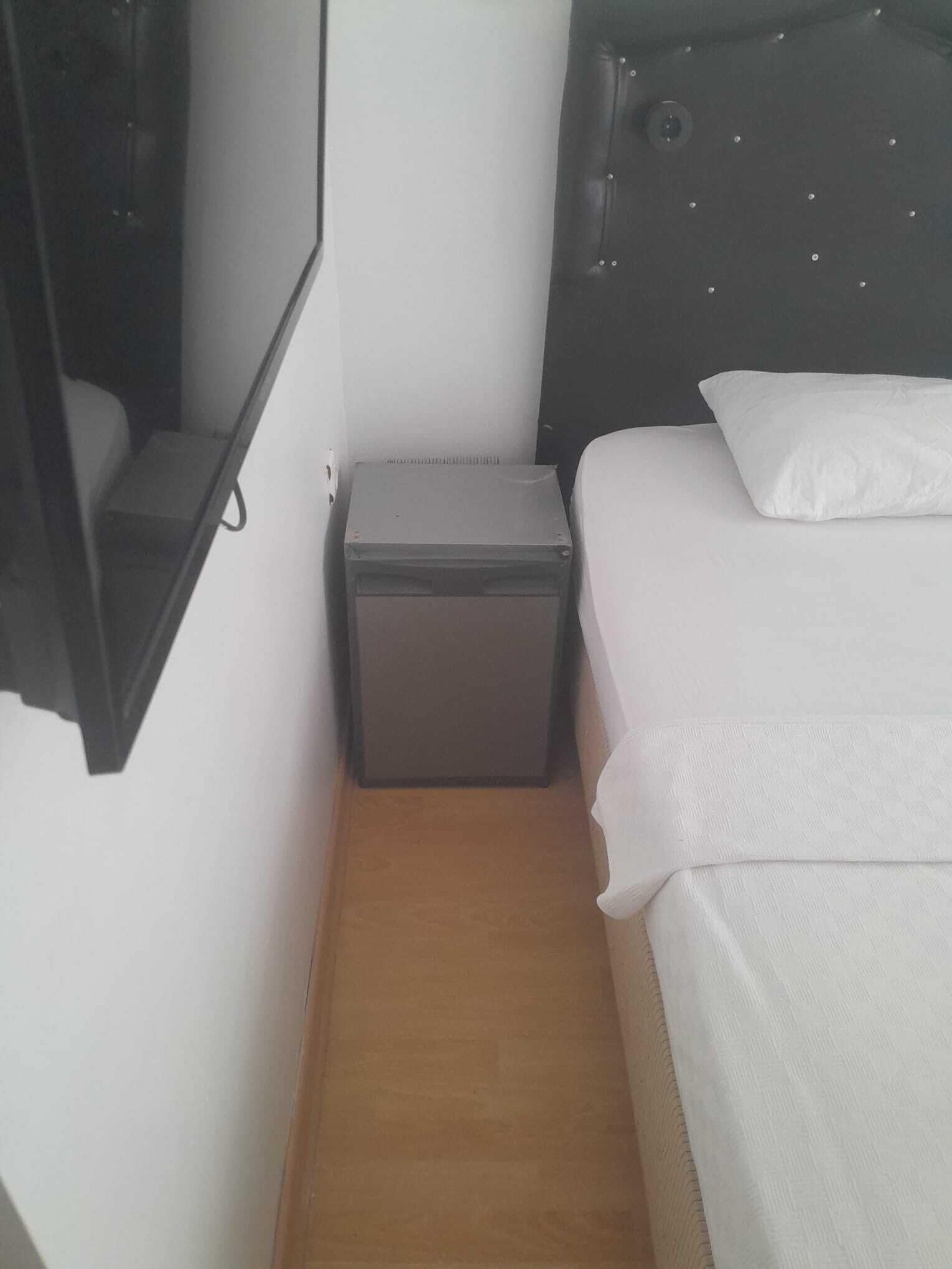 Una habitación pequeña con cama, mesita de noche, televisor montado en la pared y un pequeño cofre.