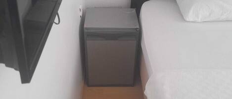 Una habitación pequeña con cama, mesita de noche, televisor montado en la pared y un pequeño cofre.