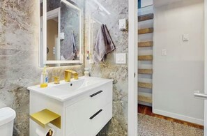 Ein Badezimmer mit Marmorwand, einem weißen Waschbecken mit goldfarbenen Armaturen, einem Spiegel und einem Handtuch an der Wand.