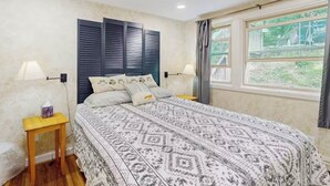 Un dormitorio con una colcha estampada, dos mesitas de noche, una mesita auxiliar, una ventana con cortinas y vistas a zonas verdes.