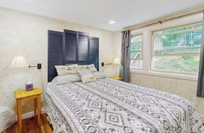 Ein Schlafzimmer mit einem gemusterten Bettbezug, zwei Nachttischen, einem Fenster mit Vorhängen und Blick auf Grünfläche.