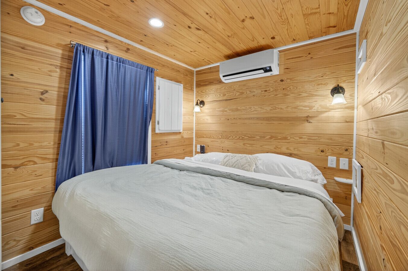 Une pièce aux murs lambrissés, comprenant un lit, un appareil de climatisation, une fenêtre avec des rideaux et une lampe fixée au mur.
