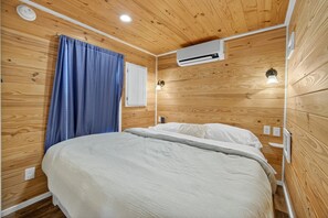 Une pièce aux murs lambrissés, comprenant un lit, un appareil de climatisation, une fenêtre avec des rideaux et une lampe fixée au mur.