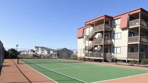 A tennis court with a net, surrounded by a brick walkway, and adjacent to a multi-story building with balconies. - NEWLY LISTED! Oceanfront and BREATHTAKING views! 3 bedrooms (North Myrtle Beach)