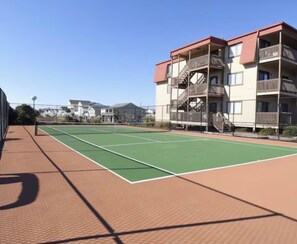 Ein Tennisplatz mit Netz, umgeben von einem Ziegelweg, neben einem mehrstöckigen Gebäude mit Balkonen.