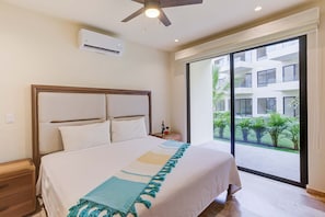 Une chambre d’hôtel avec un lit, un ventilateur de plafond, un appareil de climatisation, une table de chevet avec une bouteille et des verres, et une vue sur un immeuble et la verdure environnante.