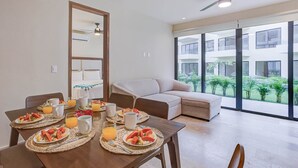 A modern living room with a dining table set for breakfast, a sofa, and a large window with a view of greenery. - Pacific coast retreat w/ kayak, bikes & pool (Tondoroque)